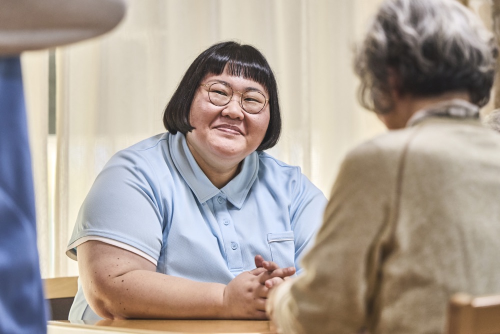戸田伊月を演じる安藤なつ。入居者と向かい合い、やわらかく微笑む。
