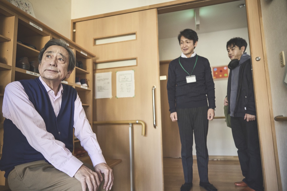 場面写真：施設の玄関で靴箱の前に腰かける豊と、戸口に立つスタッフと優輝。